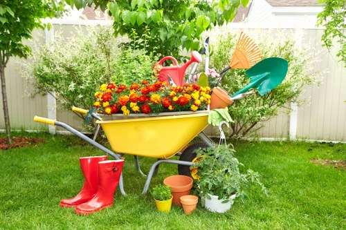A vibrant garden scene featuring a neatly maintained grassy lawn with rich green blades, bordered by a white wooden fence and lush leafy trees providing partial shade. In the centre, a yellow garden wheelbarrow is filled with bright, multicoloured flowering plants, sitting on a slightly worn paved area that transitions into the lawn. Surrounding the wheelbarrow are various gardening tools and accessories, including a red watering can, a pair of red rubber boots, and small terracotta and plastic flower pots placed on the grass and paved surface. A white potted shrub with green foliage is positioned beside the wheelbarrow, with garden hand tools submerged within or leaning against it. The backdrop features a row of well-kept shrubs and trees, creating a natural boundary, and the overall scene appears to be outdoors on a bright, mild day, ideal for gardening tasks and outdoor maintenance typical for gardens in the Neasden area, aligned with professional garden boundary care services offered by Hedge Trimming Neasden.